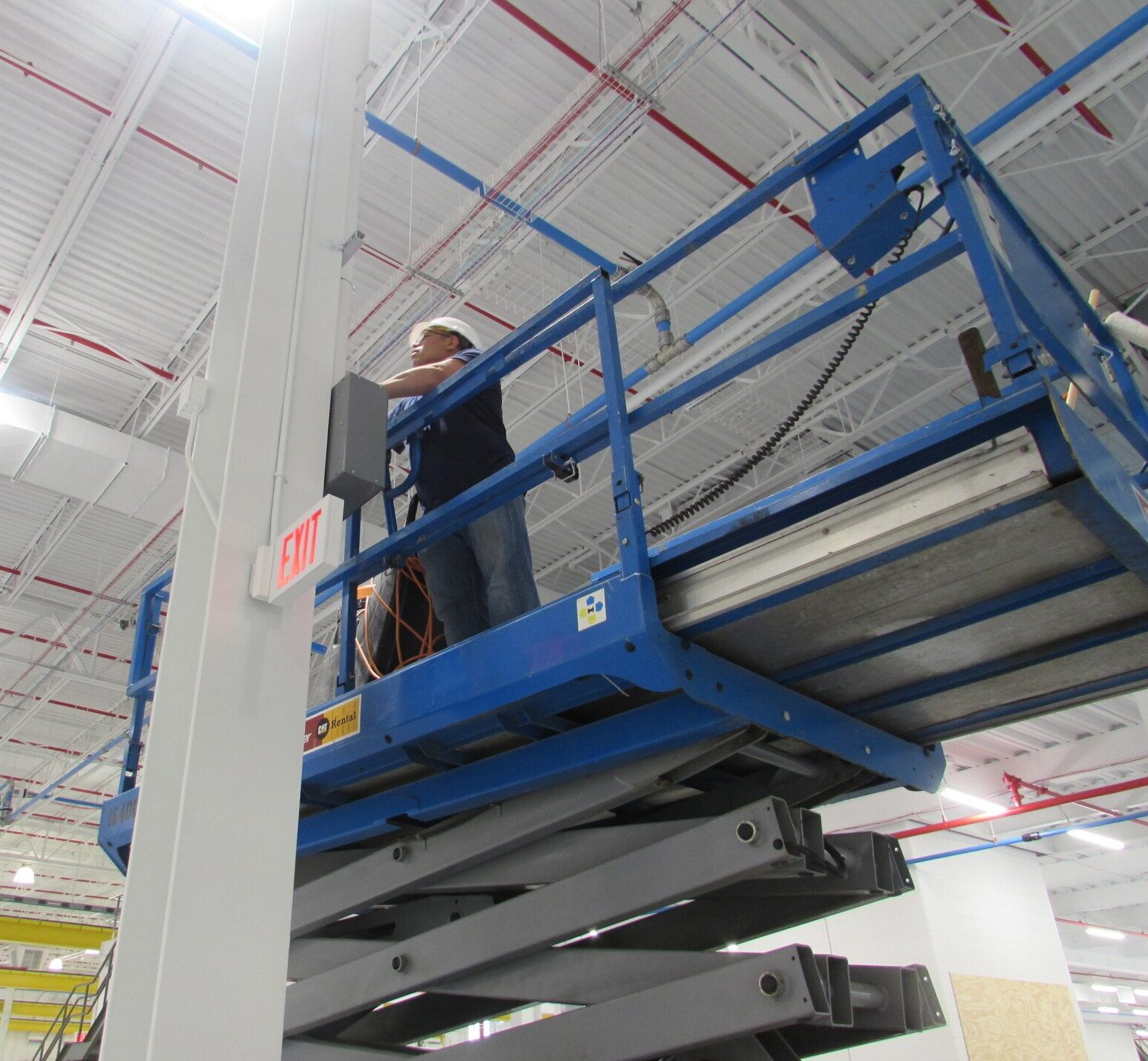 Worker on a scissor lift, wearing a hard hat, near an exit sign in a commercial building.